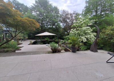 Een rustige tuin met geplaveide paden, weelderig groen en een modern beeldhouwwerk. Een parasol biedt schaduw over een zitgedeelte. Verschillende planten en bomen omringen het tafereel onder een gedeeltelijk bewolkte lucht. Gemaakt door de werkverzetter