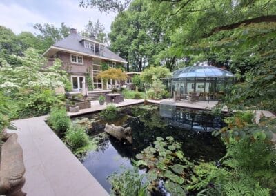 Een prachtig huis met een bakstenen buitenkant is omgeven door weelderig groen. Op de voorgrond is er een serene vijver met waterlelies en een glazen koepelstructuur ernaast, genesteld tussen hoge bomen. Buitenmeubilair is zichtbaar bij de vijver. Gemaakt door de werkverzetter