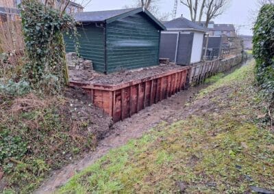 Een smalle sloot met modderig water loopt langs een kleine dijk. Boven de dijk staat een groen-witte schuur. Kale bomen en een hek vormen de achtergrond, met stukken groen en overwoekerde klimop aan de linkerkant. Gemaakt door de werkverzetter