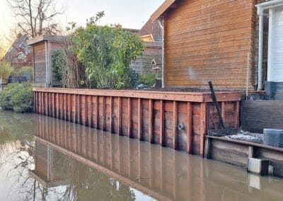 Een houten dek met een reling strekt zich uit over een kalme waterweg naast een huis. Een paar planten en struiken zijn op het dek geplaatst. Het tafereel is vredig met heldere reflecties in het water en een serene, natuurlijke sfeer. Gemaakt door de werkverzetter