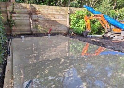 Een vers gestorte betonplaat staat in een tuin en reflecteert de omringende bomen en het zonlicht. Een kleine oranje graafmachine met een blauw zeil staat in de buurt geparkeerd. Een houten hek loopt langs de achterkant, omzoomd door groen gebladerte. Gemaakt door de werkverzetter