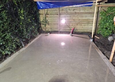 Een vers gelegde betonnen plaat droogt onder kunstlicht met houten omheining en groen op de achtergrond. Een blauw zeil bedekt een deel van het gebied. Er blijft een kleine hoeveelheid water op het oppervlak achter. Gemaakt door de werkverzetter