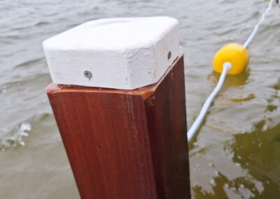 Een houten paal met een witte bovenkant steekt uit in een watermassa, verbonden door een ketting aan een rij boeien met gele drijvers. Het water is lichtjes choppy en de kustlijn is zichtbaar op de achtergrond onder een bewolkte lucht. Gemaakt door de werkverzetter