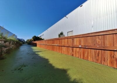 Een smal kanaal bedekt met groene algen loopt langs een metalen pakhuis. Een houten hek scheidt het kanaal van het gebouw, onder een helderblauwe lucht. Spaarzaam groen omzoomt de linkerkant van het kanaal. Gemaakt door de werkverzetter