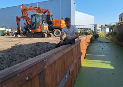Een bouwvakker meet een houten constructie naast een groen pad in een buitenruimte. Een oranje graafmachine en industriële gebouwen zijn zichtbaar op de achtergrond onder een helderblauwe lucht. Gemaakt door de werkverzetter