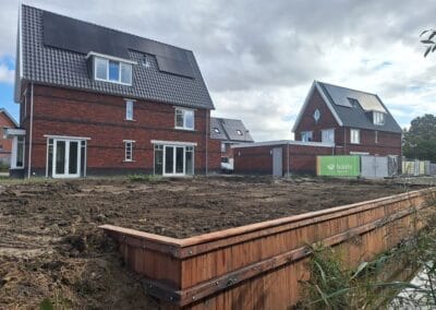 Twee moderne bakstenen huizen met zonnepanelen en grote ramen staan naast elkaar, grenzend aan een houten talud en een stukje kale aarde. Een groen bord van een bouwbedrijf is zichtbaar. Bewolkte lucht op de achtergrond. Gemaakt door de werkverzetter