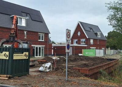 Een bouwplaats in een woonwijk met twee bakstenen huizen met zonnepanelen. Er staat een graafmachine en een grote container voor. Er is een snelheidslimiet en een bord met verboden te parkeren zichtbaar. Er liggen grond en bouwmaterialen verspreid. Gemaakt door de werkverzetter