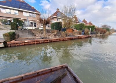 Uitzicht op een woonwaterkant met verschillende huizen met zonnepanelen. Een houten dek en keermuur zijn zichtbaar langs de waterkant, met een gedeeltelijk bewolkte lucht en kalm water dat de omgeving weerspiegelt. Gemaakt door de werkverzetter
