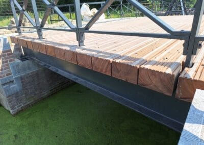 Een houten voetgangersbrug met metalen leuningen strekt zich uit over een groene, met algen bedekte waterweg. Het oppervlak van de brug bestaat uit netjes uitgelijnde houten planken. Aan één kant van de brugsteunen is metselwerk zichtbaar. Op de achtergrond zijn bomen te zien. Gemaakt door de werkverzetter