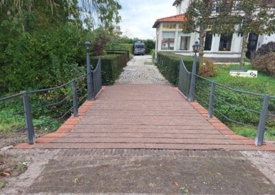 Een kleine houten brug met leuningen leidt naar een huis met een rood pannendak. De brug overspant een grindpad en het huis is omgeven door groen. Er staat een voertuig geparkeerd op de oprit. Gemaakt door de werkverzetter