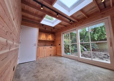 Een moderne houten kamer met dakramen en grote glazen deuren. Het interieur heeft houten muren en plafond, een betonnen vloer en een kitchenette met kasten. Buiten is er groen zichtbaar door de glazen deuren. Gemaakt door de werkverzetter