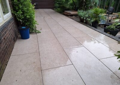 Een schoon, betegeld terras met grote beige tegels, gedeeltelijk nat van de regen. Planten en struiken grenzen aan het gebied, met een blauwe emmer aan de zijkant en een kleine kas zichtbaar op de achtergrond. Gemaakt door de werkverzetter