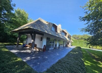 Een charmant huis met een rieten dak en grote ramen, gelegen te midden van weelderig groen. Het heeft een ruim stenen terras, een houten bank onder een overhang en is omgeven door verzorgde heggen en bomen onder een helderblauwe lucht. Gemaakt door de werkverzetter