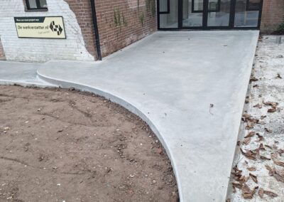 Een nieuw betonnen trottoir buigt om de hoek van een bakstenen gebouw. Gevallen bladeren liggen verspreid en er is een klein tuintje zichtbaar naast het pad. Er staat een geel waarschuwingsbord bij de ingang. Gemaakt door de werkverzetter