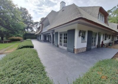 Een ruim huis met een rieten dak, omgeven door weelderig groen. Het heeft grote ramen, houten luiken en een breed stenen terras. De omgeving is vredig, met bomen en heggen die het tafereel omlijsten. Gemaakt door de werkverzetter