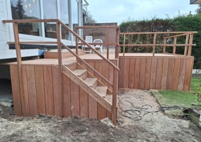 Een nieuw gebouwd houten terras met trap en leuningen is bevestigd aan een huis. Het terras kijkt uit op een tuin met gras, aarde en een gesnoeide heg. Een verlengsnoer is zichtbaar op de grond bij het terras. Gemaakt door de werkverzetter