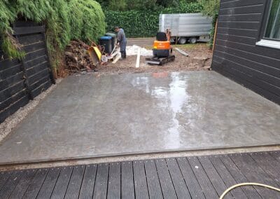 Een vers gestorte betonplaat naast een zwarte houten constructie is zichtbaar. Een persoon op de achtergrond werkt in de buurt van bouwmaterialen en een kleine graafmachine. Weelderig groen omringt het gebied.