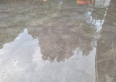 Een plas water op een betonnen oppervlak reflecteert bomen en structuren, wat een vervormd spiegeleffect creëert. De oppervlaktetextuur en lichte rimpelingen zijn zichtbaar, wat de reflecterende kwaliteit van de scène versterkt.