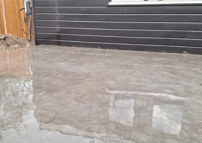 Een vers gestorte betonnen oprit die droogt voor een modern huis met donkere horizontale gevelbekleding. Links is een raam met gesloten jaloezieën zichtbaar en rechts piept een houten hek met planten.