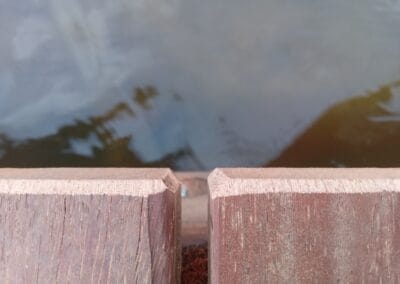 Close-up van een opening tussen twee houten planken met water eronder zichtbaar. De planken hebben een ruwe textuur en het water reflecteert licht, wat een kalme, serene sfeer creëert.