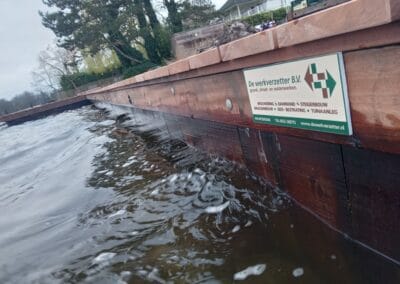 Een close-up van een houten steiger met een bord voor "De werkverzetter B.V." gedeeltelijk onder water. Bomen en een gebouw zijn zichtbaar op de achtergrond. Het water lijkt lichtjes choppy, wat duidt op beweging of wind.