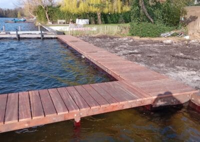 Een houten steiger strekt zich uit over een kalm waterlichaam met een haakse bocht. Een met gras begroeide oever en bomen, sommige met felgeel blad, zijn op de achtergrond. Een paar boten liggen afgemeerd bij de verre kustlijn.