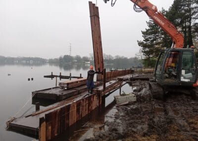 Een bouwplaats bij een meer waar een arbeider een grote stalen plaat begeleidt die door een oranje graafmachine wordt opgetild. Het gebied is modderig en op de achtergrond zijn houten constructies en bomen zichtbaar onder een bewolkte lucht.