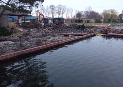 Een bouwplaats bij een waterlichaam, met graafmachines, puin en een gedeeltelijk gebouwde constructie. Een kleine boot ligt in de buurt aangemeerd. Bomen en gebouwen zijn zichtbaar op de achtergrond onder een bewolkte lucht.