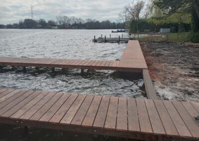 Een houten pier strekt zich uit over een kalm meer met bewolkte luchten. Het water kabbelt zachtjes tegen de constructie. Rondom de pier staan bomen en een met gras begroeide oever. Op de achtergrond is nog een dok zichtbaar.