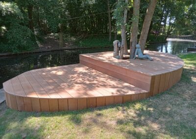 Houten dek bij een kanaal met ingebouwde zitplaatsen rond een boom. Een sculptuur van twee abstracte figuren staat op het dek. Het gebied is omgeven door weelderige groene bomen en gras, en zonlicht werpt speelse schaduwen op het hout.