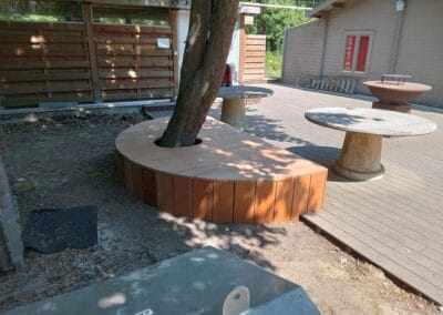 Een houten dek met ronde tafels en een boom omgeven door een ronde houten bank. Een houten hek en gebouw staan op de achtergrond en zonlicht werpt gevlekte schaduwen op het tafereel.