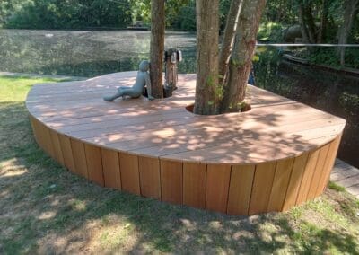 Een houten dek omringt een boom, gelegen aan een meer. Een grijze, mensachtige figuur zit leunend tegen de boom. De omgeving is kalm en pittoresk met weelderig groen en water op de achtergrond.