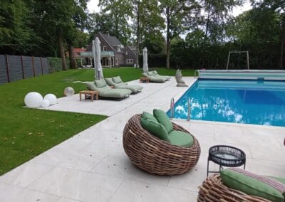 Een achtertuin met een zwembad omgeven door groen. Ligstoelen met parasols staan op het terras bij het zwembad. Een rieten stoel met groene kussens staat op de voorgrond en een huis is zichtbaar op de achtergrond tussen de bomen.