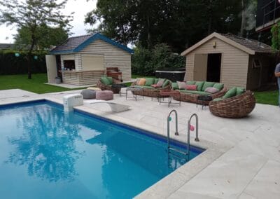 Een achtertuin met een zwembad en meubilair aan het zwembad, inclusief stoelen met kussens en banken. Er zijn twee houten schuurtjes, groen en bomen rondom het gebied, wat een ontspannende buitenruimte creëert.