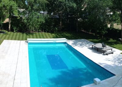 Breed uitzicht op een achtertuin met een groot rechthoekig zwembad omgeven door netjes gemaaid gras. Een klein schuurtje staat rechts en op de achtergrond staat een huis tussen de bomen, met terrasstoelen en een tafel bij het zwembad.