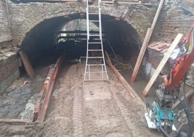 Een gedeeltelijk overstroomde bouwplaats met een stenen boog. Een metalen ladder leunt tegen de boog en houten balken worden gebruikt ter ondersteuning. Machines en slangen zijn zichtbaar en de grond lijkt modderig.