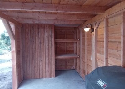 Een houten schuurinterieur met een onafgewerkte vloer, met ingebouwde planken aan de achterwand. Een aan de muur gemonteerde lamp zorgt voor licht en een grote overdekte barbecuegrill is zichtbaar in de rechter benedenhoek.