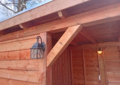 Houten schuurhoek met een metalen en glazen lantaarn aan de muur. De structuur heeft natuurlijke houten lambrisering onder een lattendaksteun. Een kleine wandlamp is binnenin zichtbaar. Bomen zijn op de achtergrond te zien tegen een bewolkte lucht.