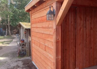 Houten schuurtje met schuin dak, met een aan de muur bevestigde lantaarn. Het schuurtje staat in een tuin met bomen en een ander kleiner schuurtje is zichtbaar op de achtergrond. De grond is een mix van aarde en beton.